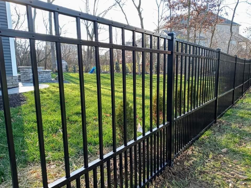 Black metal fence bordering a green lawn, with trees and a house in the background.