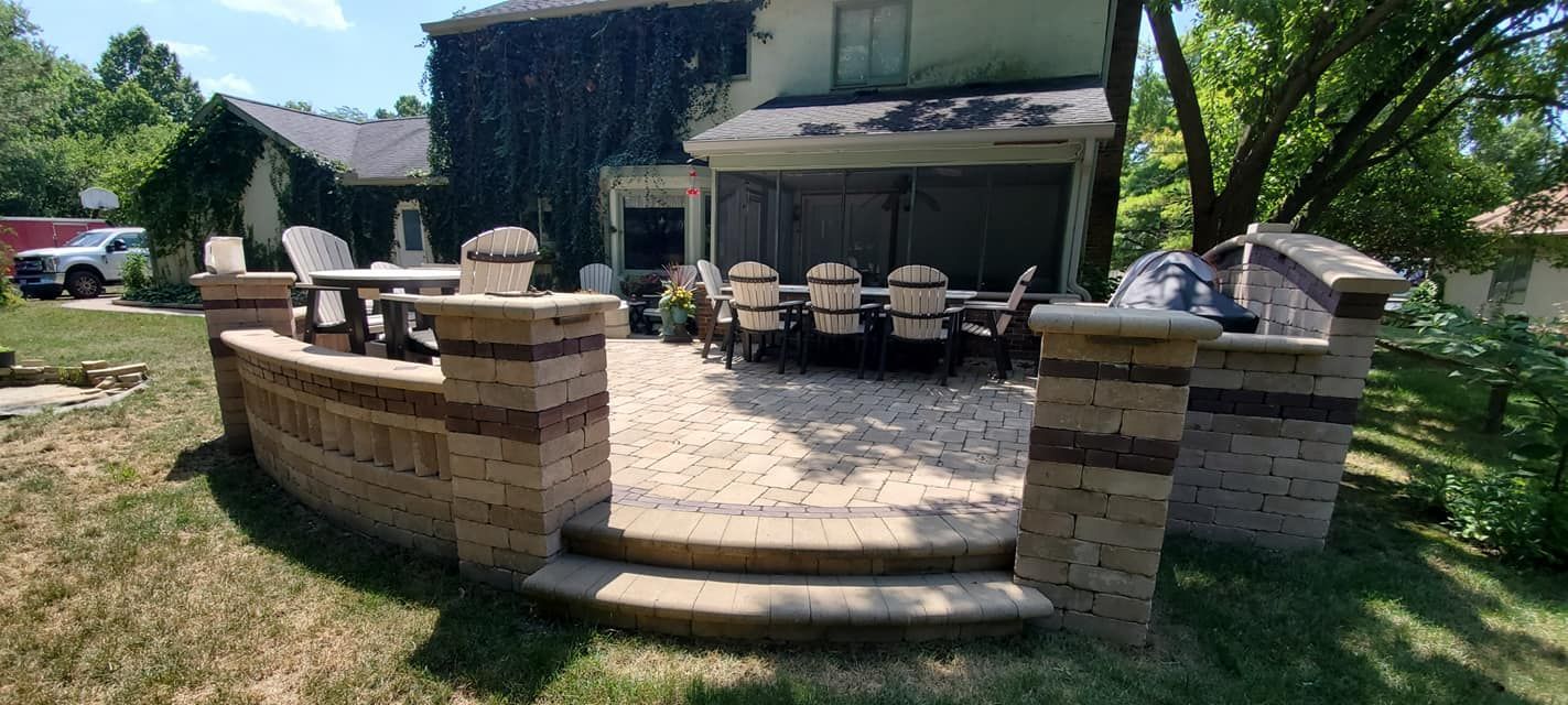 Brick patio with seating area, steps, and outdoor dining set, surrounded by grass and foliage.