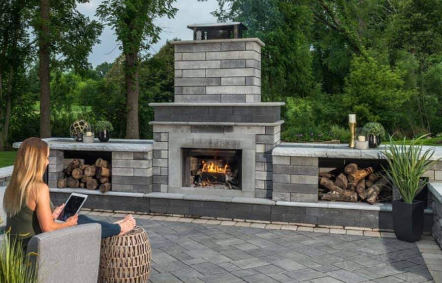Woman using tablet near a lit outdoor fireplace with firewood storage, set on a patio.