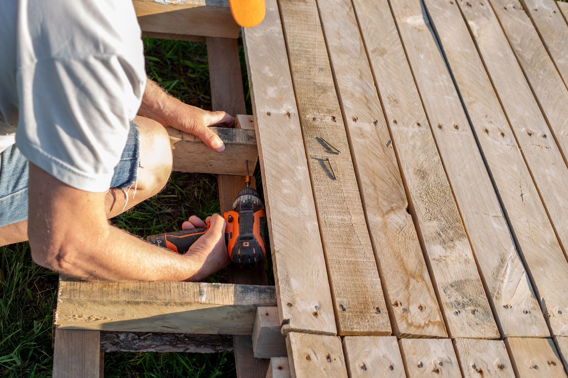 Person using a drill to attach wooden planks, building a deck outdoors.