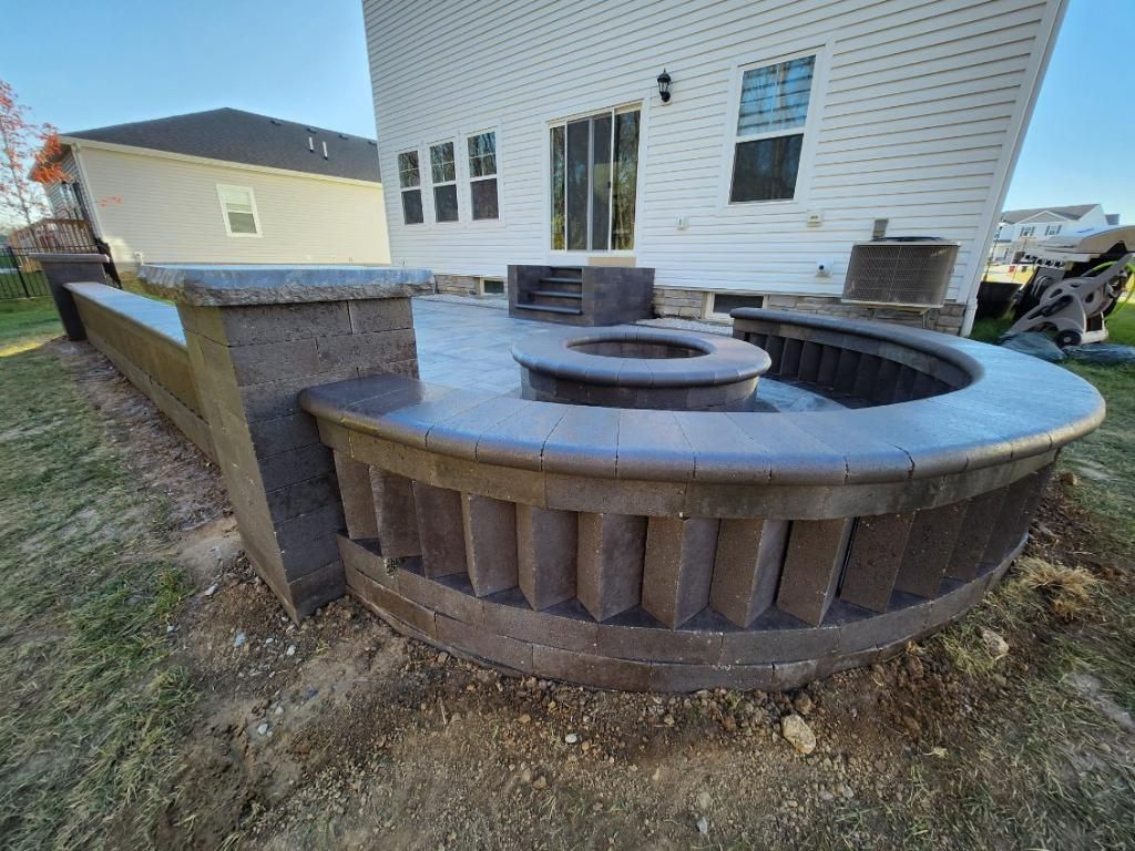Stone patio with fire pit, built-in seating, and a low retaining wall, near a white house.