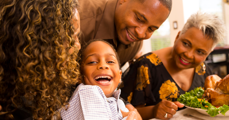 Family at a dinner table smiling; a child laughs. A plate of food is in the center.