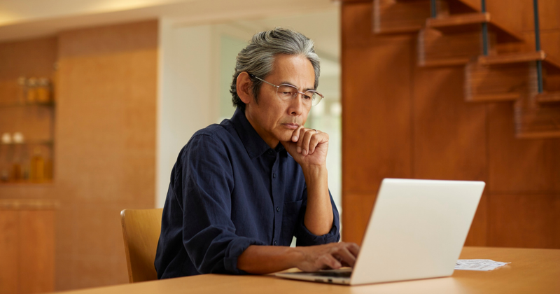 Man working on laptop, resting his chin on his hand. He is in a modern interior setting near stairs.