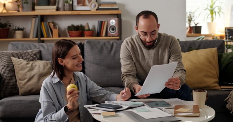 Couple reviewing documents together on a coffee table in a living room; woman eating an apple.