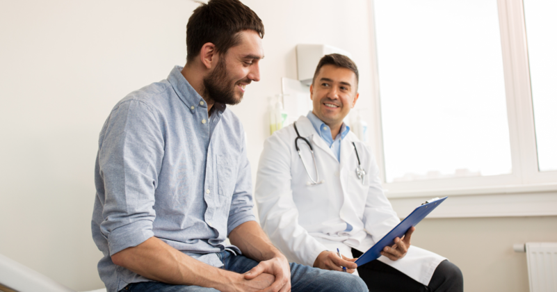 Man sitting, smiling, talking with a smiling doctor in a medical office; doctor holding a clipboard.