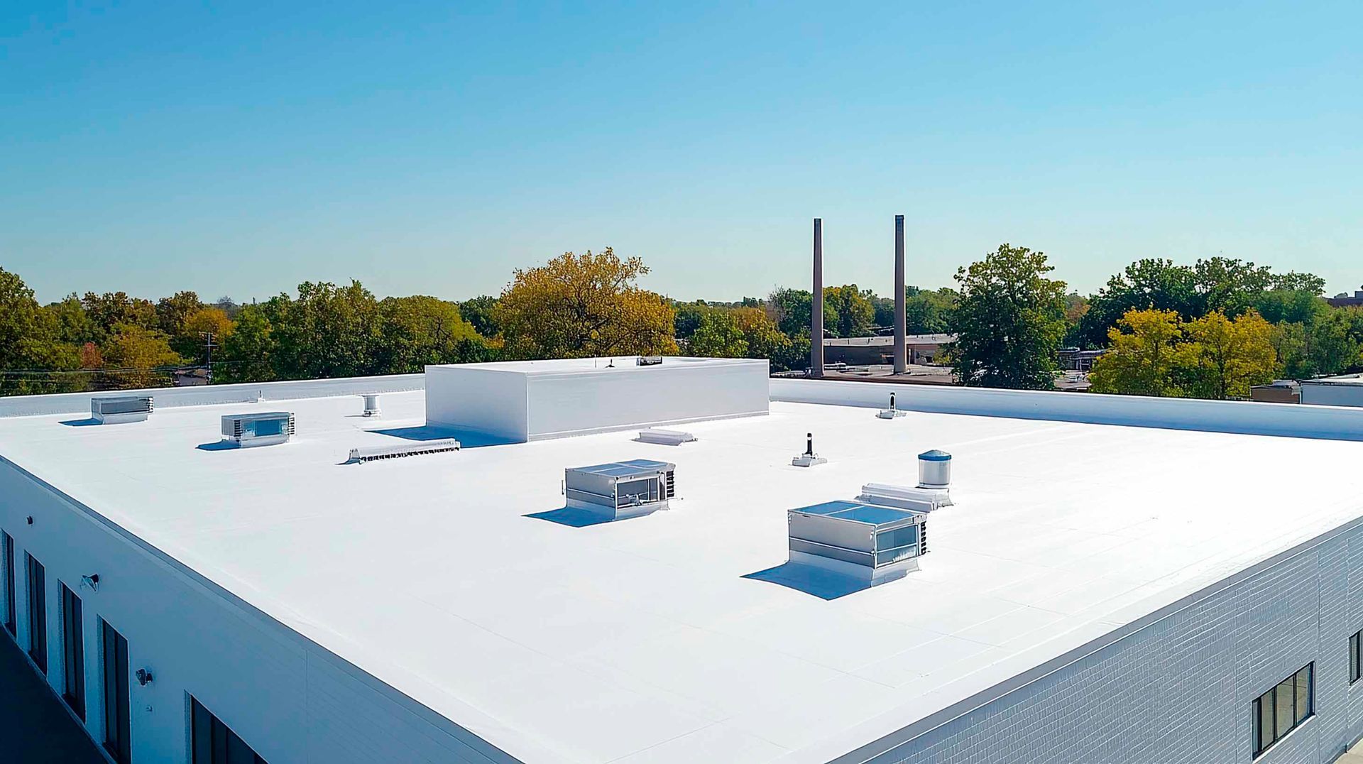 White flat commercial roof with HVAC units, trees, and smokestacks against a blue sky.