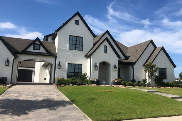 White brick house with black trim, a driveway, and green lawn under a blue sky.