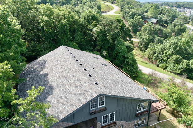 Gray shingle roof of a house surrounded by green trees, with a road in the distance.