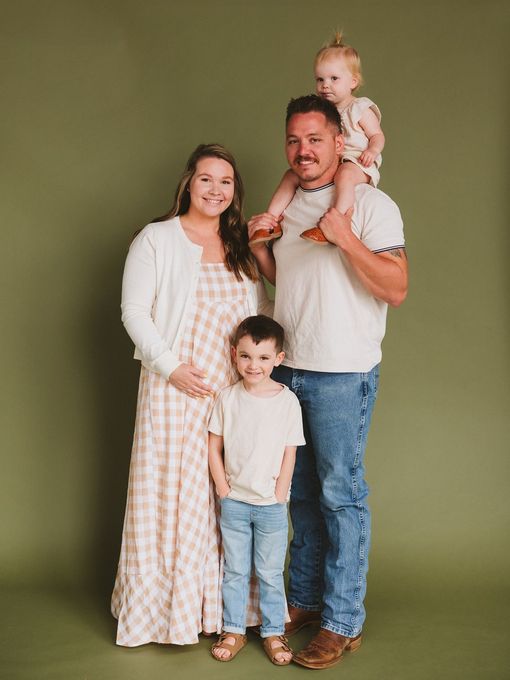 Family of four posing in front of a green backdrop. Woman in dress and cardigan, man with child on shoulders, boy standing.