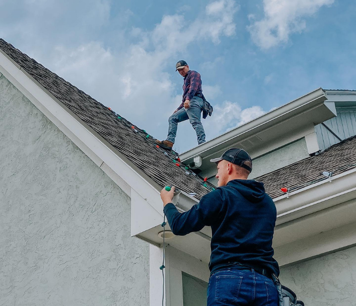 Two workers installing Christmas lights on a roof; one on the roof, one on a ladder.