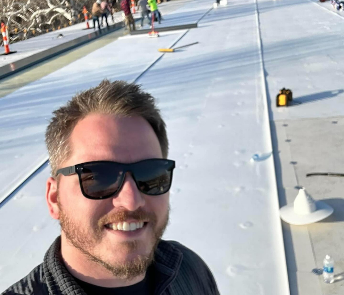 Man in sunglasses smiles, taking a selfie on a flat, white roof with construction in the background.