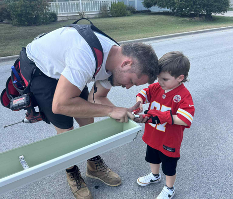Man and young boy working on a gutter outside. Man is leaning over, boy holds a tool, both focused.