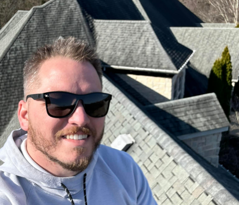 Man in sunglasses on a rooftop, smiling; gray shingles, trees visible in the background.