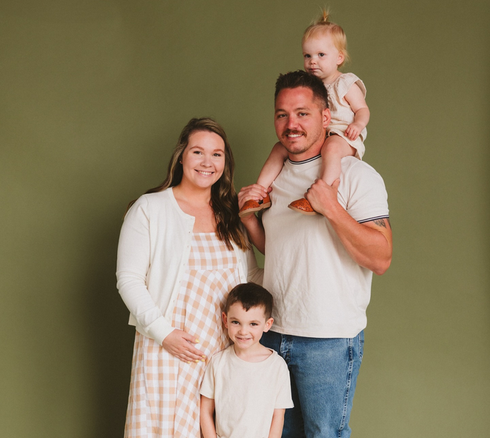 A man is holding a baby on his shoulders while standing next to a pregnant woman and two children.