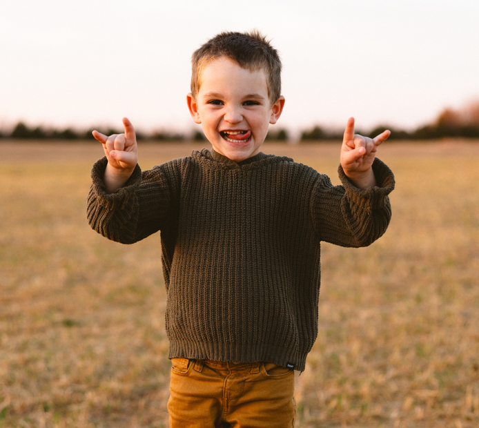 A young boy in a sweater is standing in a field making a rock and roll sign.