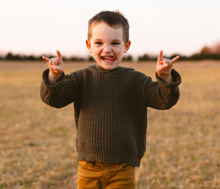 Young boy with brown hair, smiling and making a rock-and-roll hand gesture in a field, wearing a green sweater and brown pants.