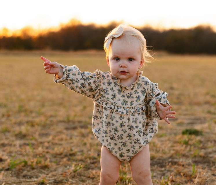 A blonde toddler in a floral bodysuit points towards the left in a field during sunset.