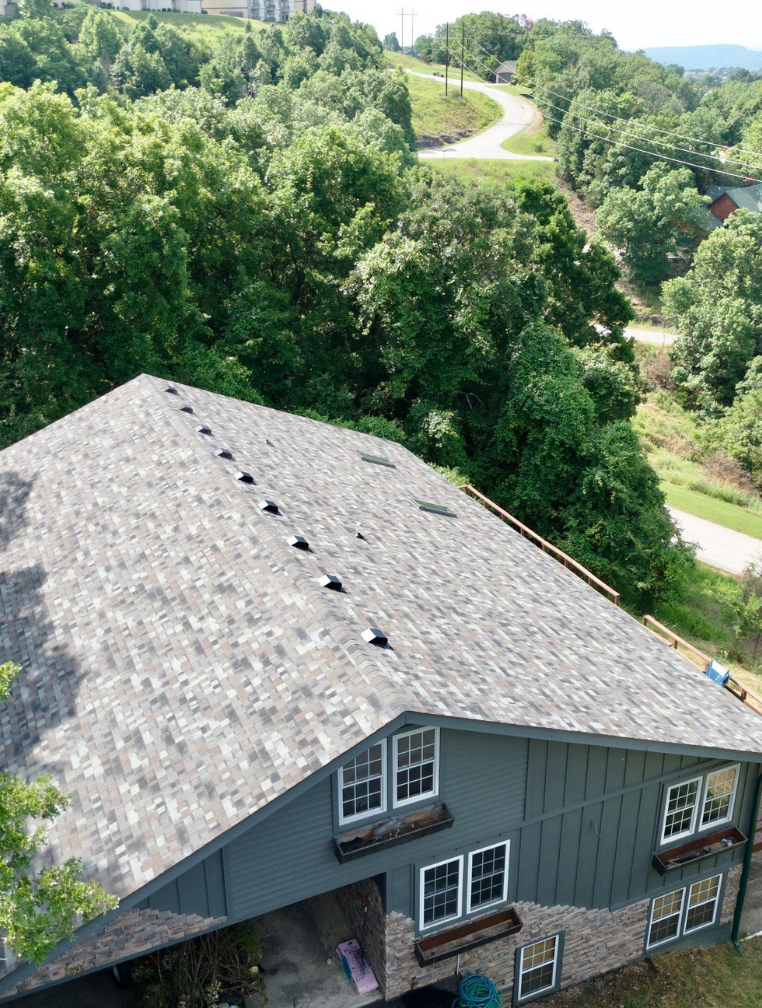 Overhead view of a house with a gray shingled roof nestled in a green, hilly, wooded landscape.