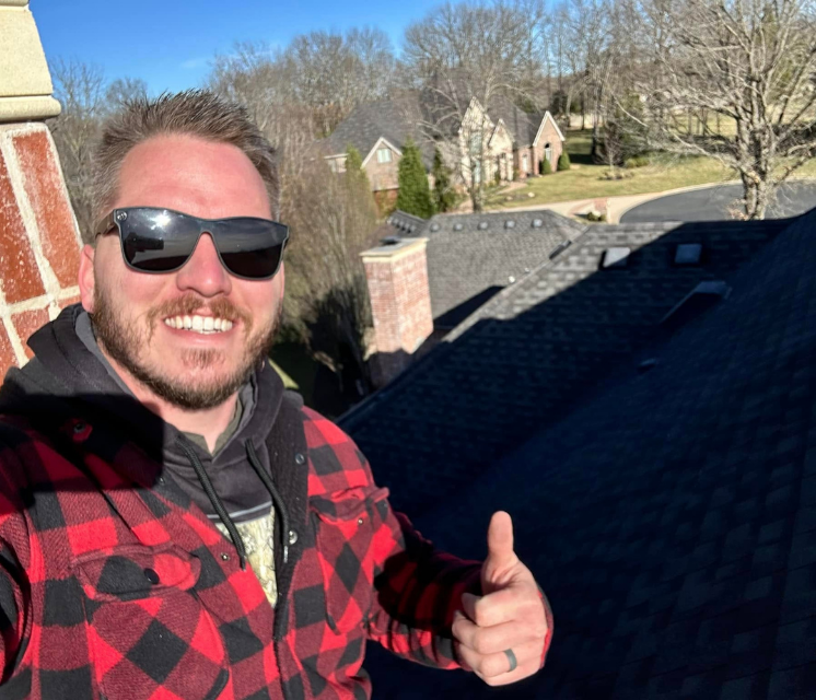 A man in a red and black plaid jacket and sunglasses smiles with a thumbs-up on a rooftop; houses and trees in the background.