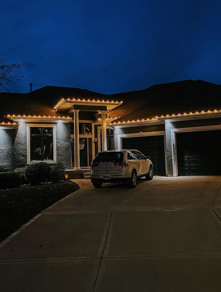 A house at dusk with string lights along the roof and a white car parked in the driveway. Dark blue sky.