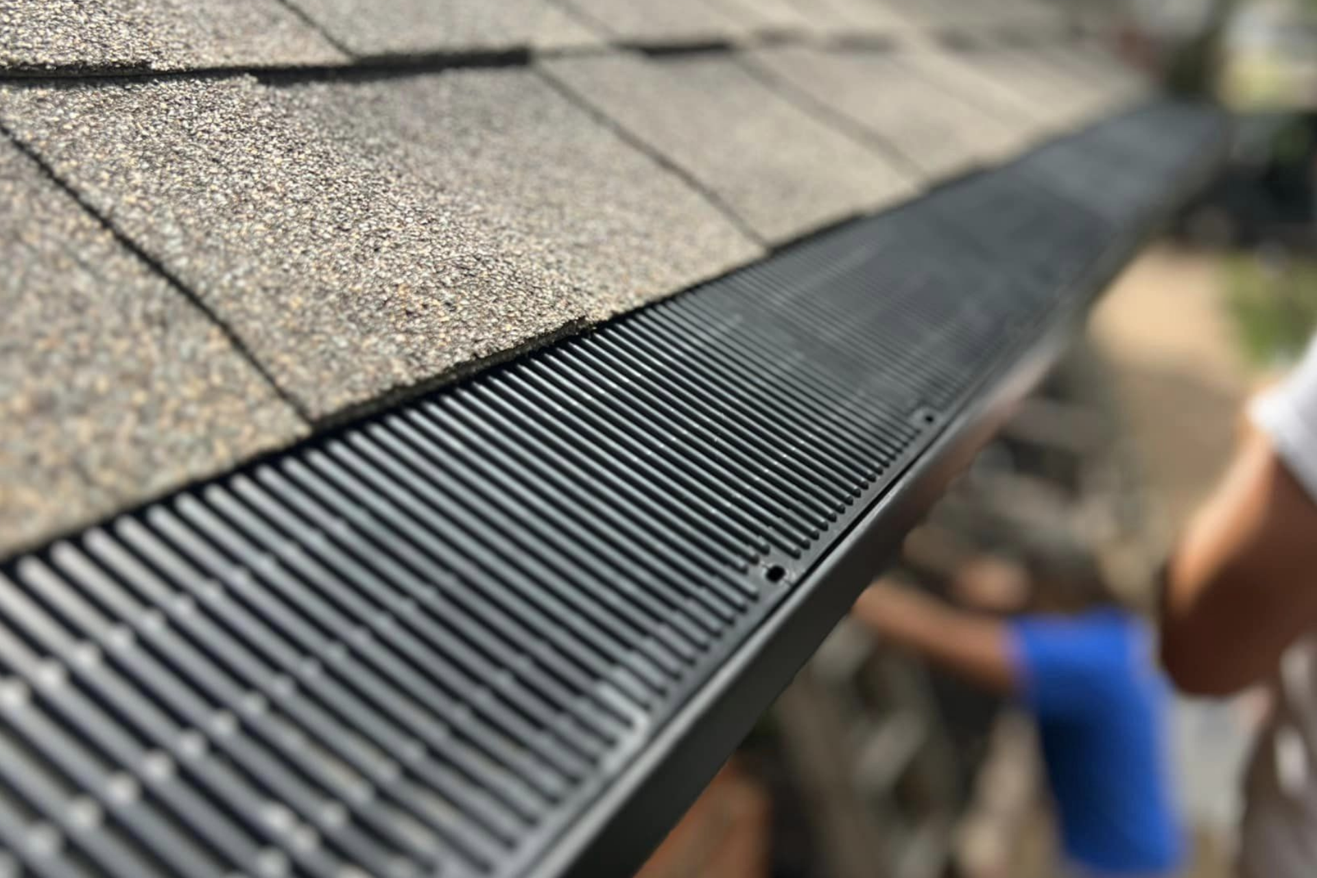 Close-up of a black gutter guard installed on a roof with gray shingles; a person is visible in the background.