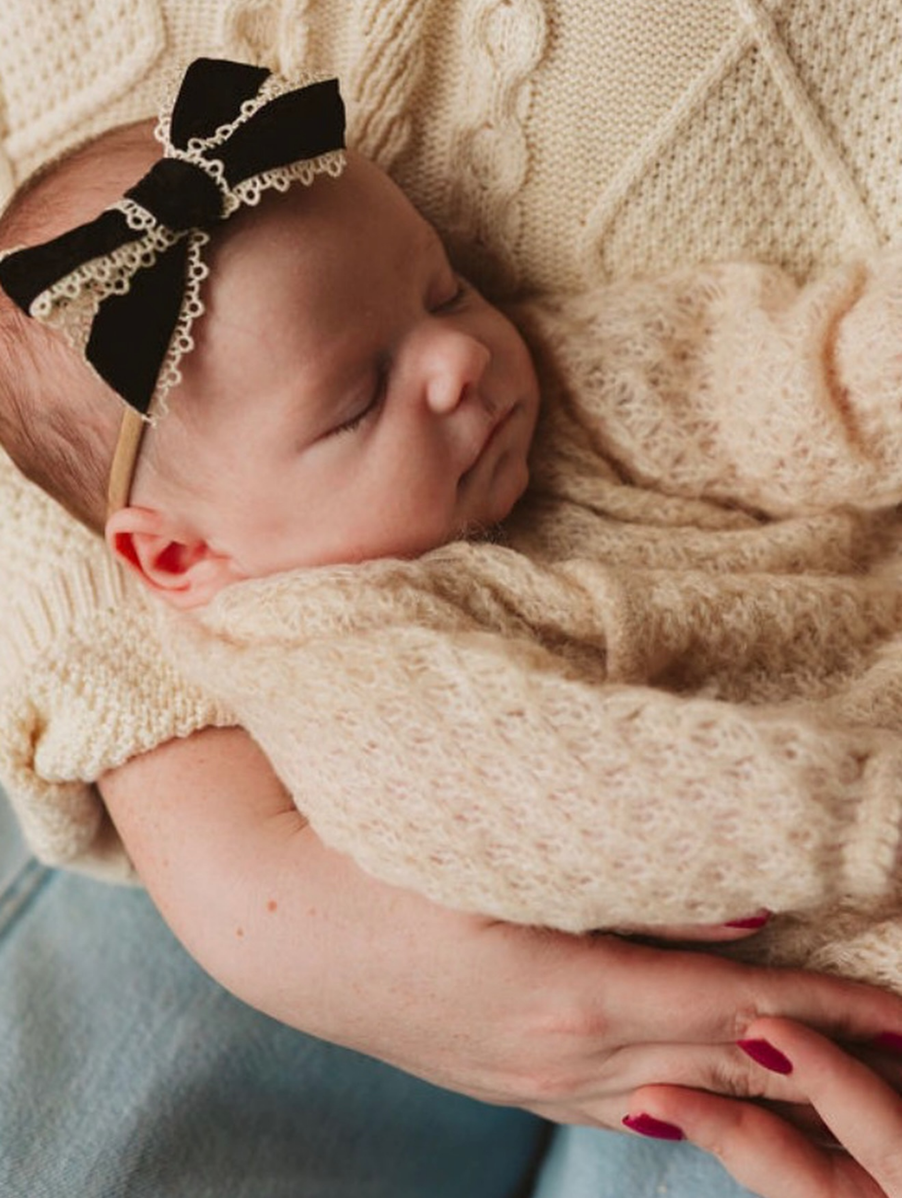Newborn sleeping in someone's arms, wearing a black bow with white trim on its head.