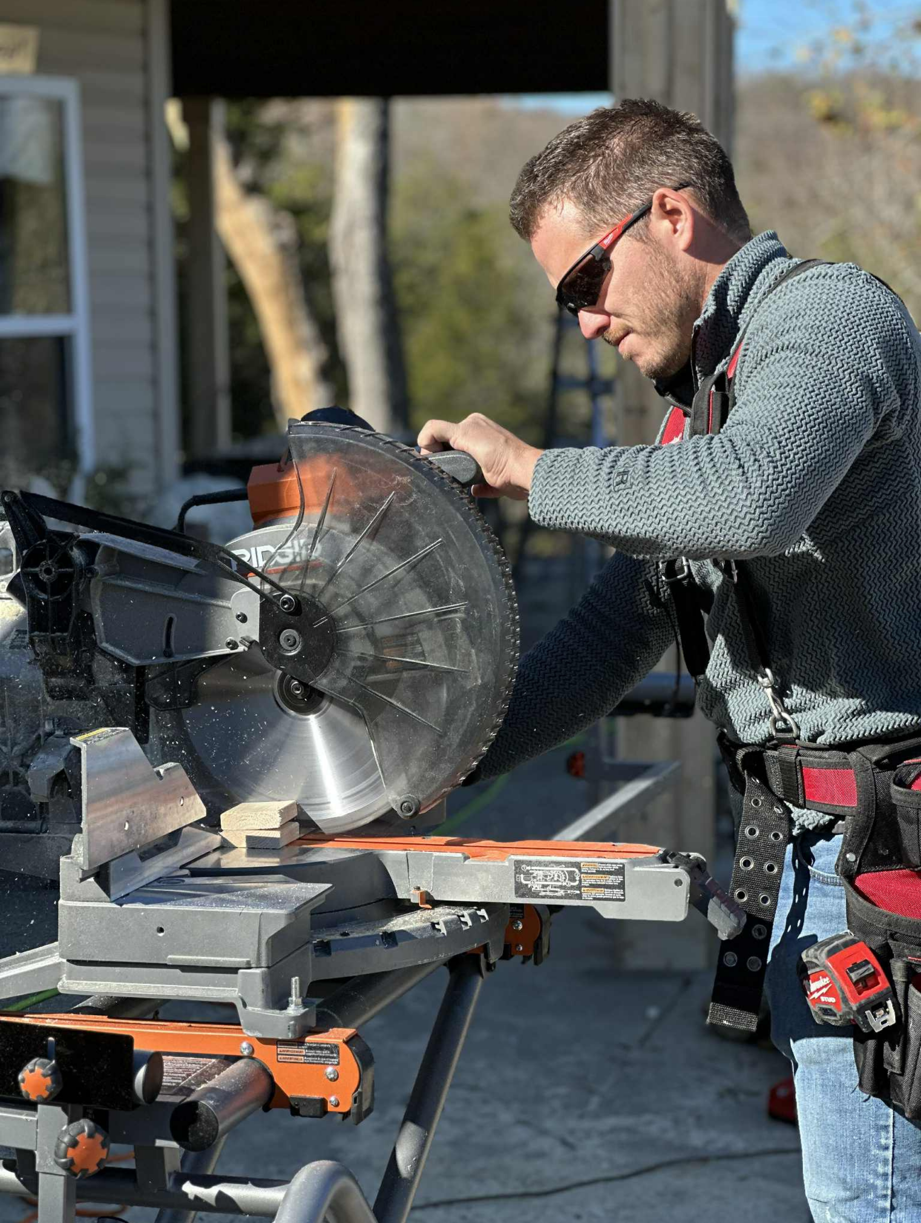 Man using a miter saw outdoors, wearing sunglasses and a tool belt, preparing to cut wood.