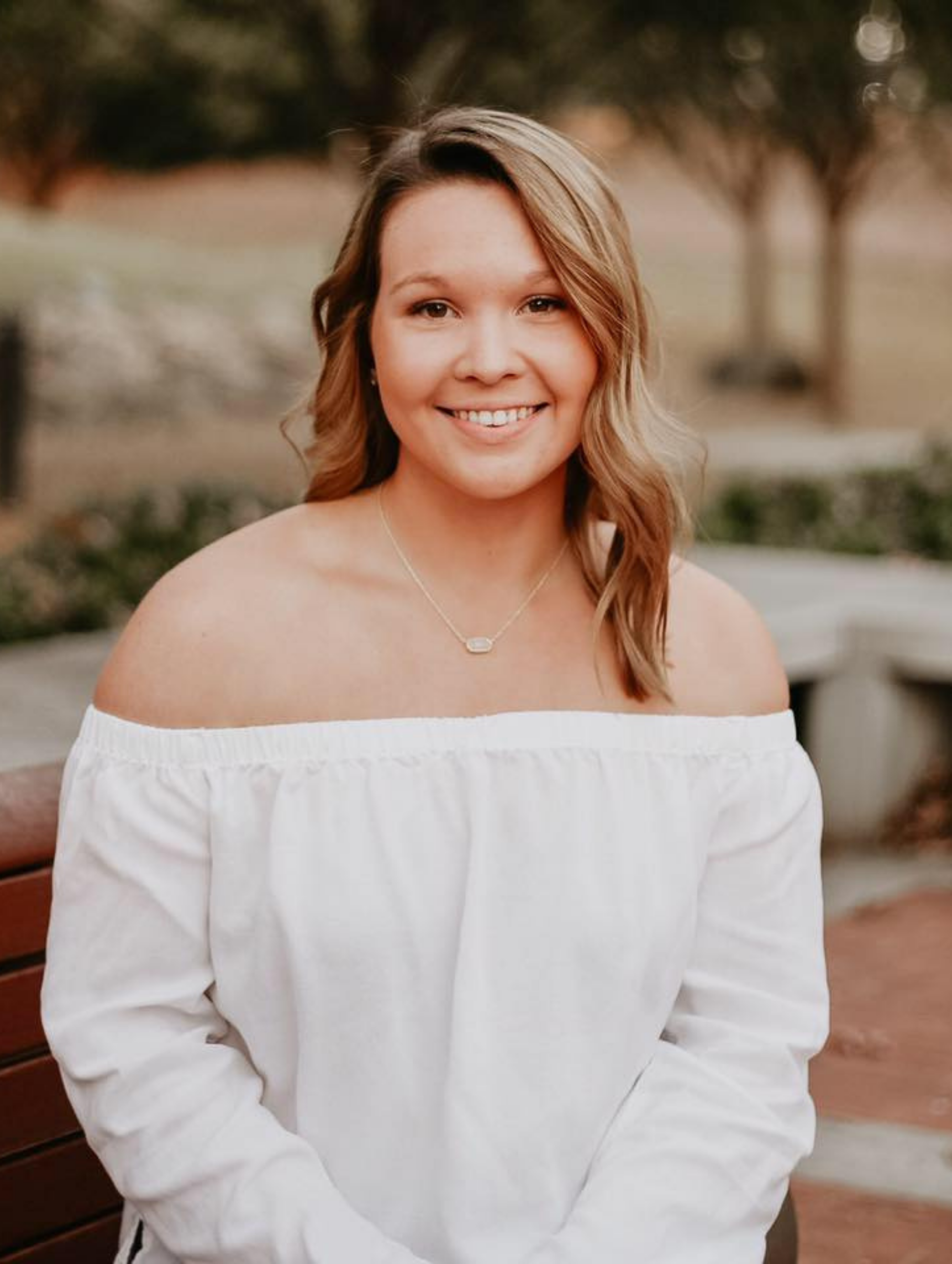 Woman with wavy blonde hair smiles, wearing a white off-the-shoulder shirt, sitting on a park bench.