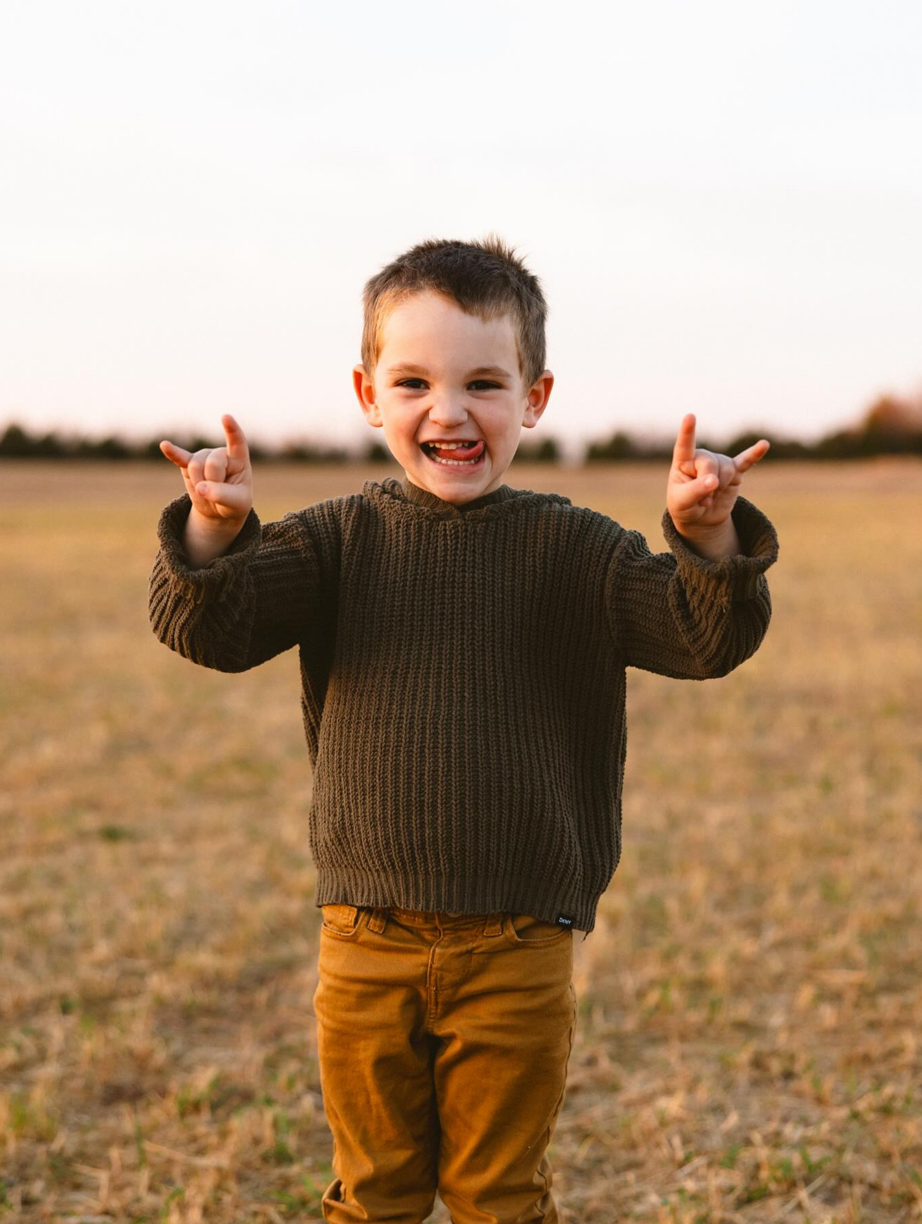 Boy in a brown sweater and pants making a hand gesture with tongue out, field background.