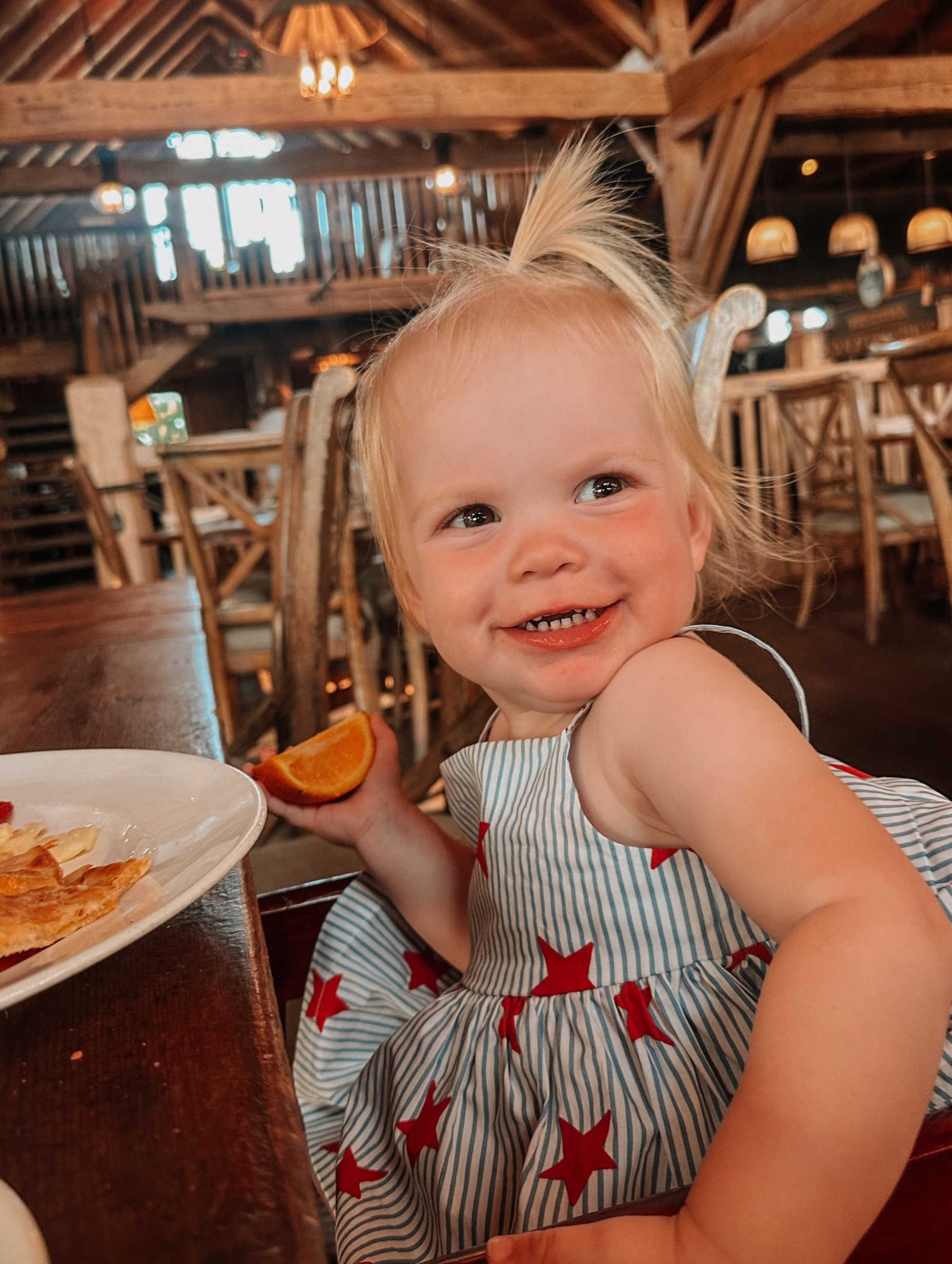 Smiling toddler holds orange slice, sitting at a table in a rustic restaurant.