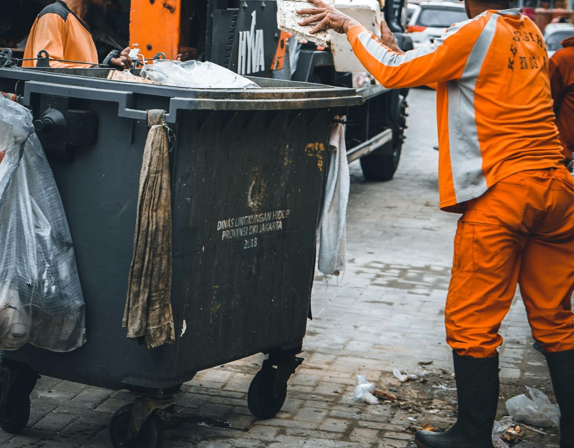A sanitation worker in an orange uniform empties a large trash bin into a truck on a paved street.
