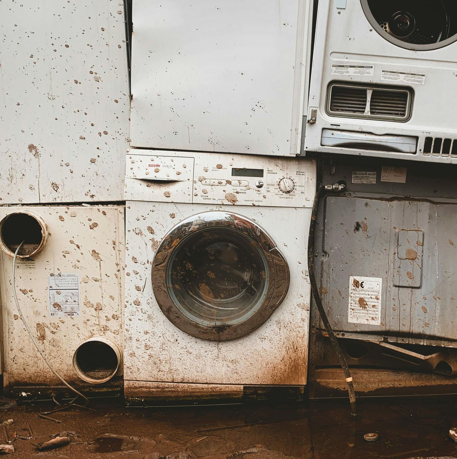 A stack of discarded, mud-caked washing machines and dryer units sitting on dark, wet ground.