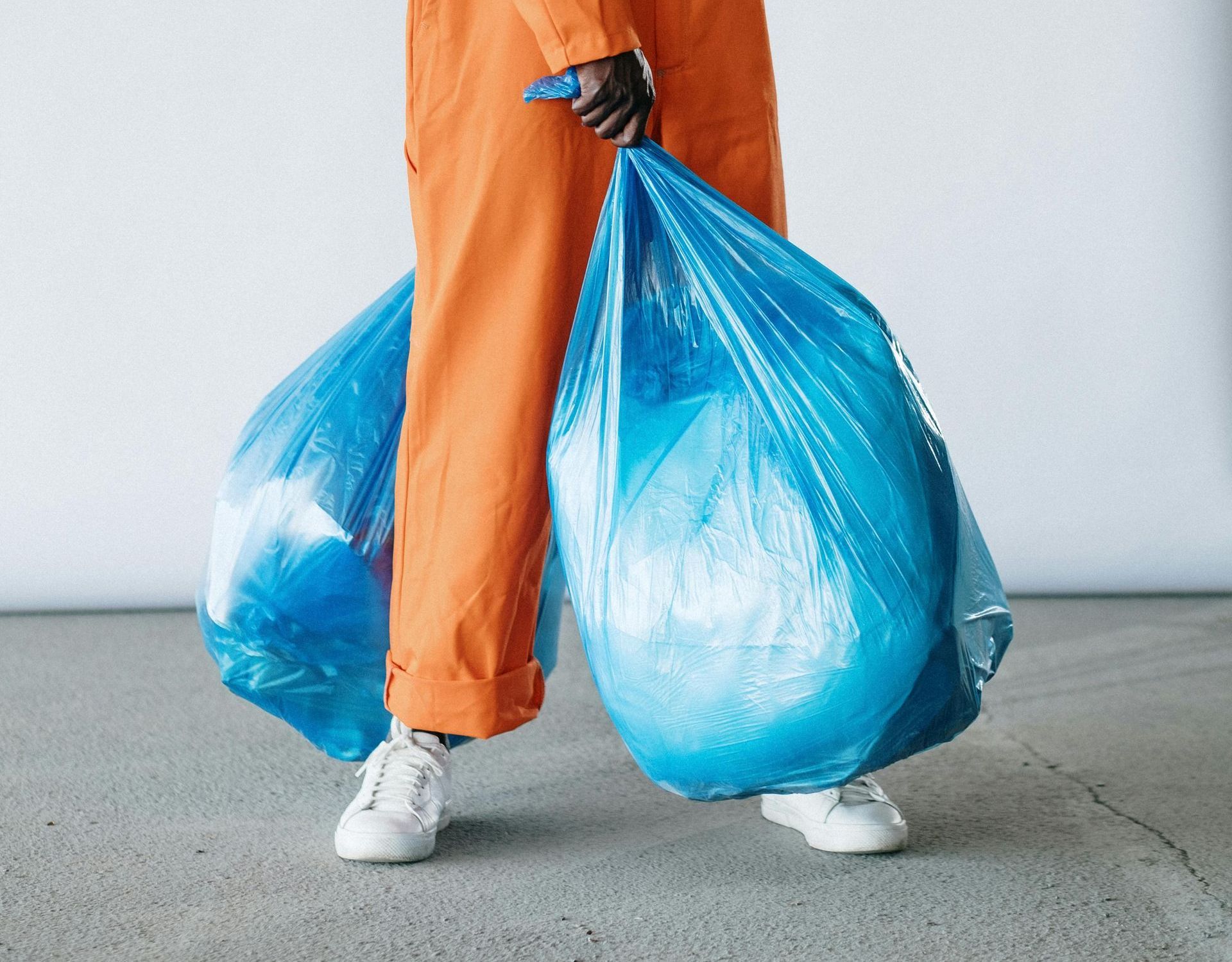A person wearing orange coveralls and white sneakers carries two large blue trash bags.