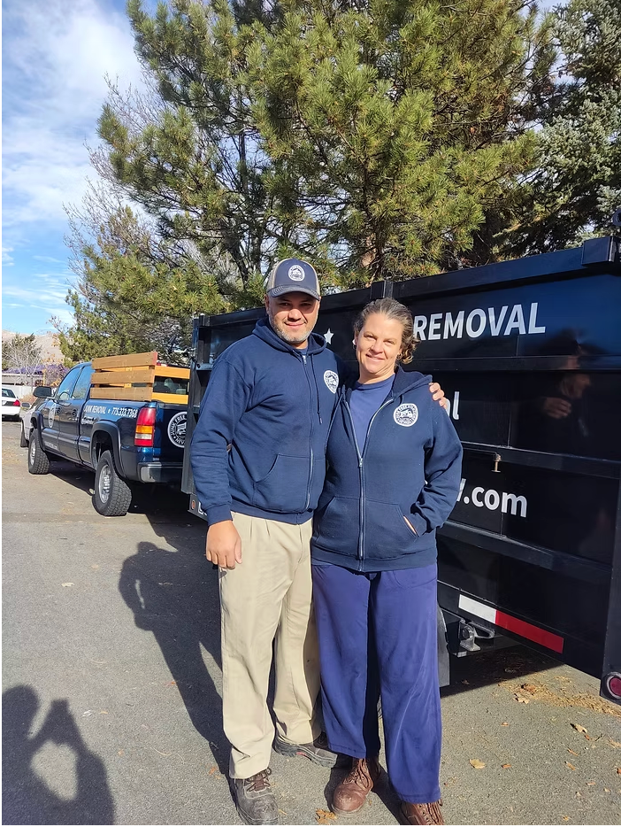 Two people in matching blue hoodies stand by a black dumpster with a truck in the background.