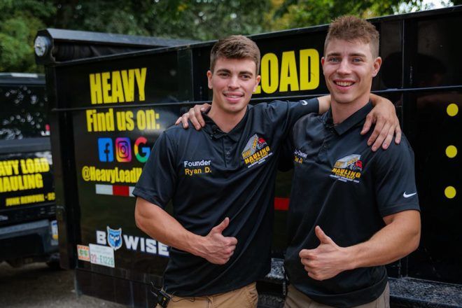 Two men with thumbs up in front of a dump truck, logo