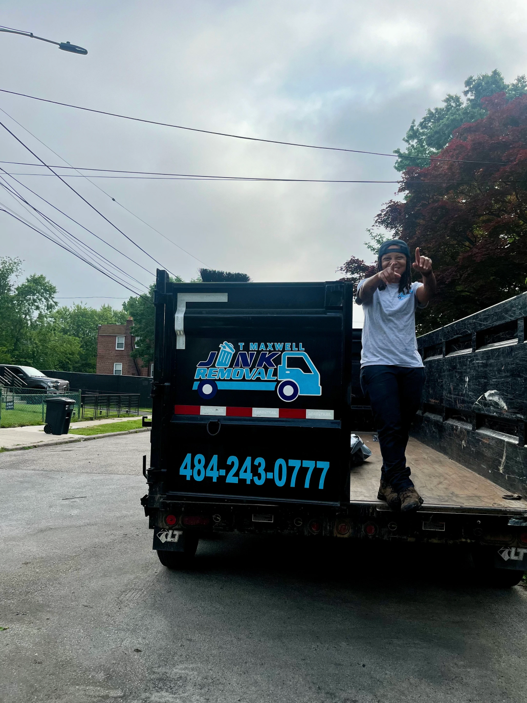 Person on a truck gives a thumbs up. A dumpster rental truck with blue graphics and phone number on the back. Outdoors.