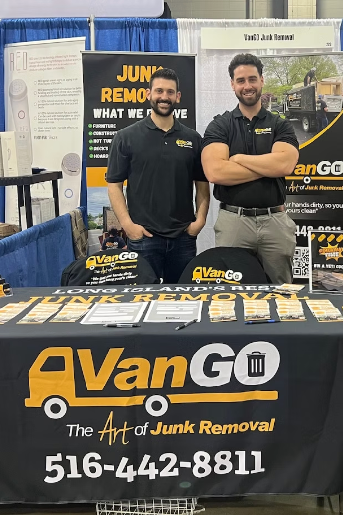 Two men at a VanGo junk removal booth. One has arms crossed. Black shirts, navy backdrop, brochures on table.