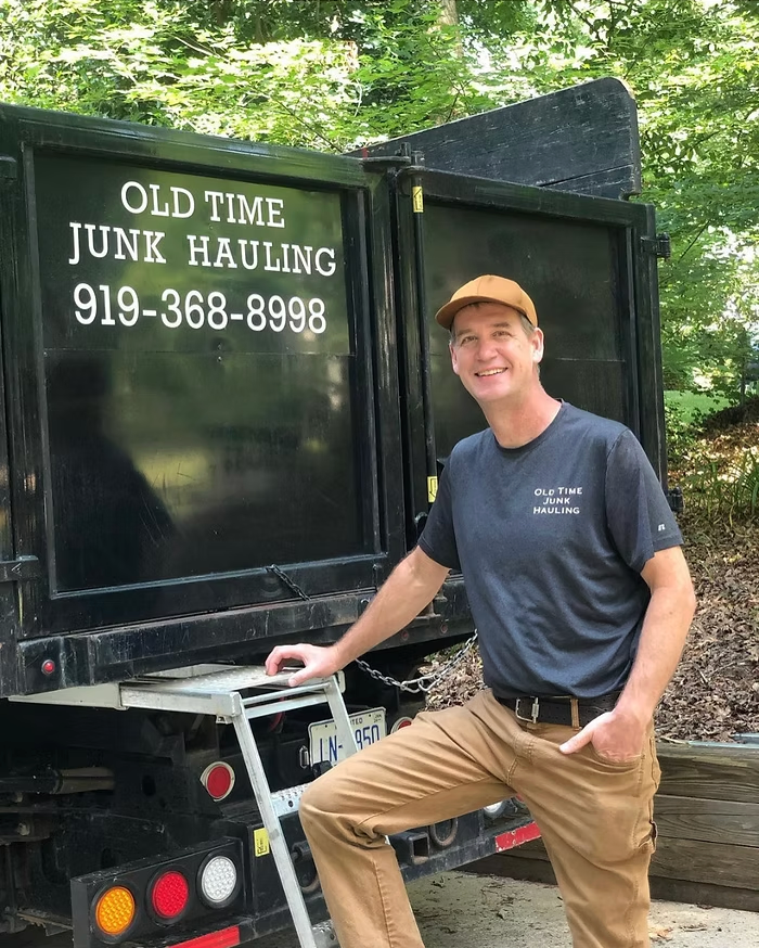 Man stands by a black junk hauling truck, smiling, wearing a hat, t-shirt, and khakis. The truck has