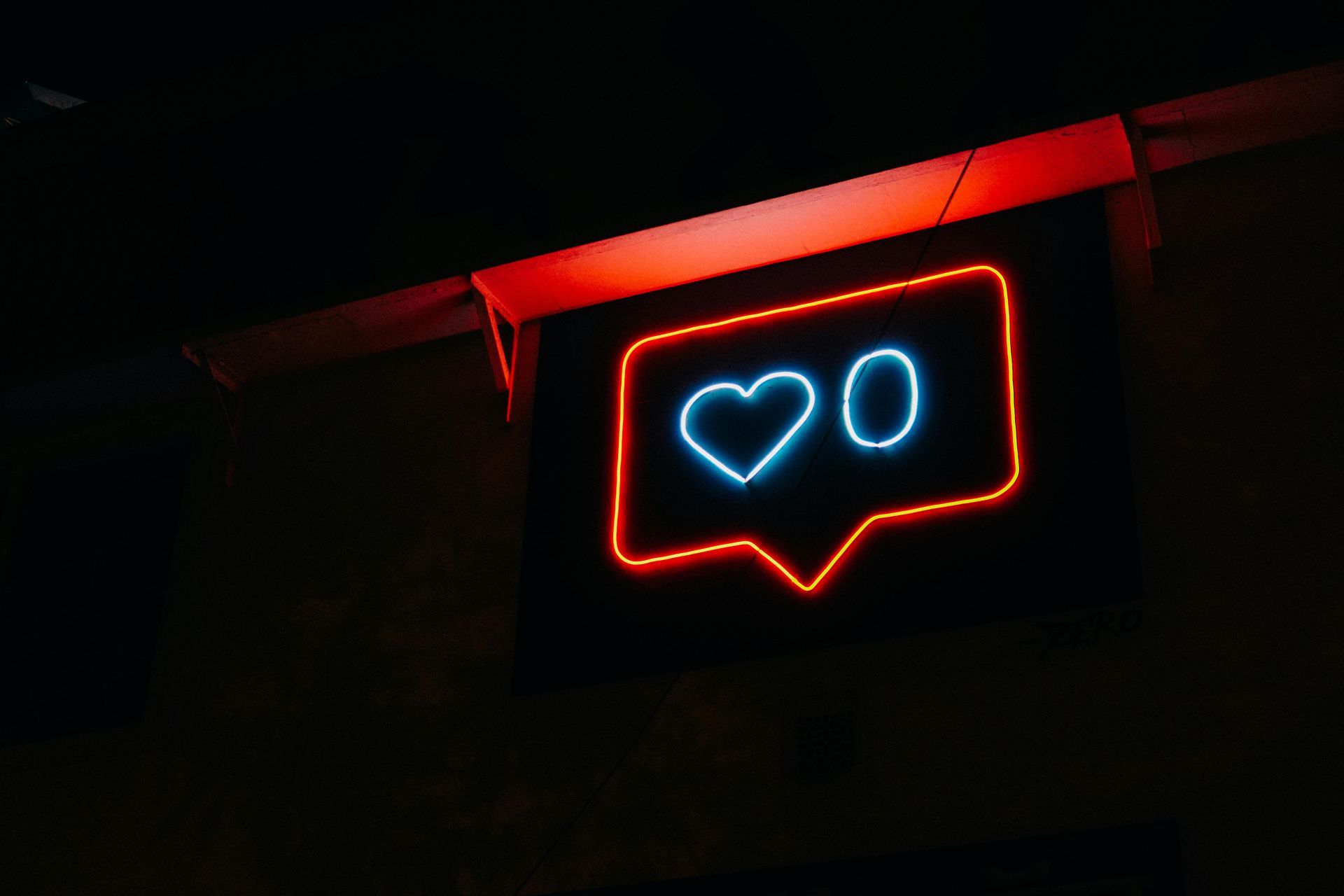 Neon sign of a heart and number 0 inside a speech bubble, glowing blue and red against a dark background.