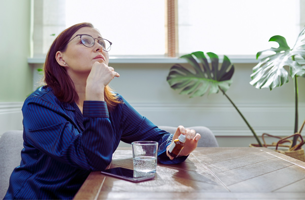 A woman wearing glasses is sitting at a table with a glass of water and a pill bottle.