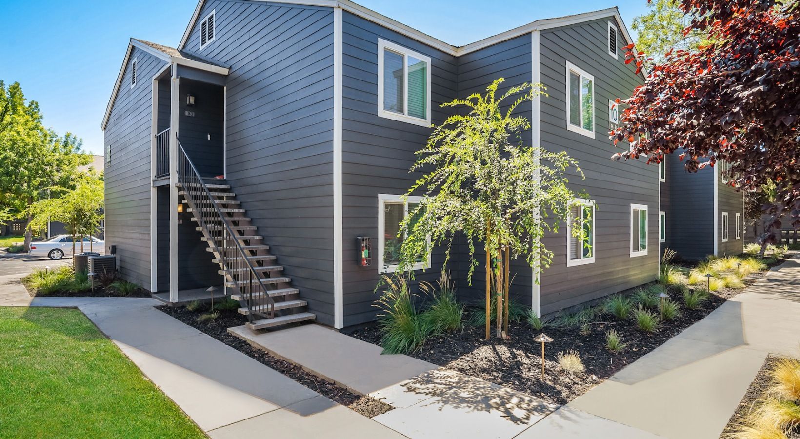 Two-story apartment building with dark gray siding, stairs, and landscaping under a clear blue sky.