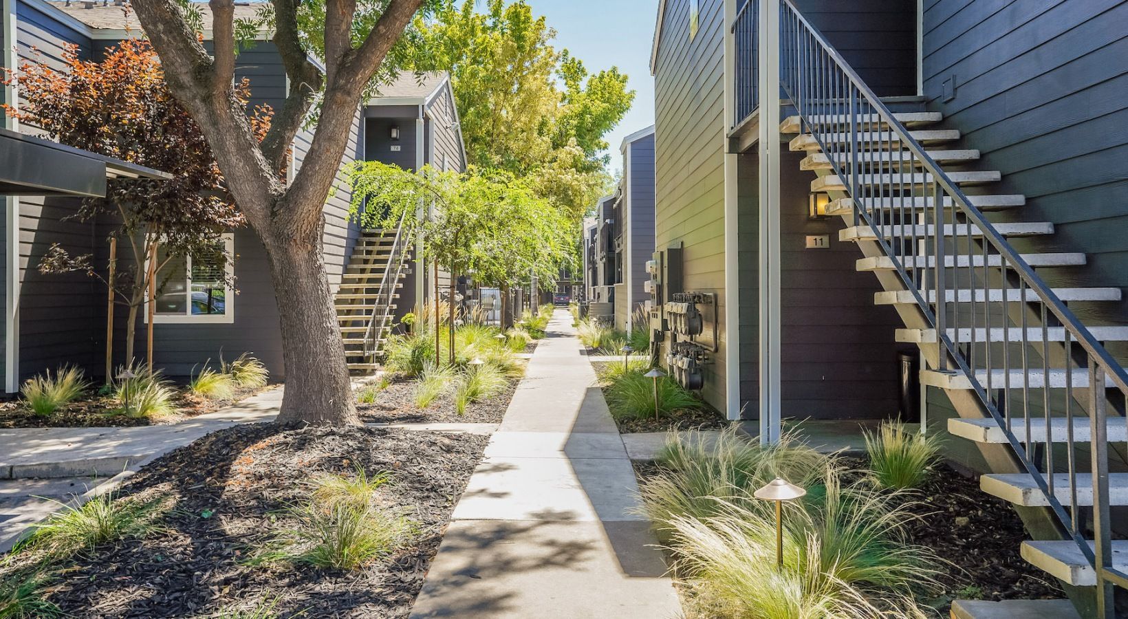 Apartment complex exterior: paved walkway, trees, dark buildings with exterior staircases, and landscaping.