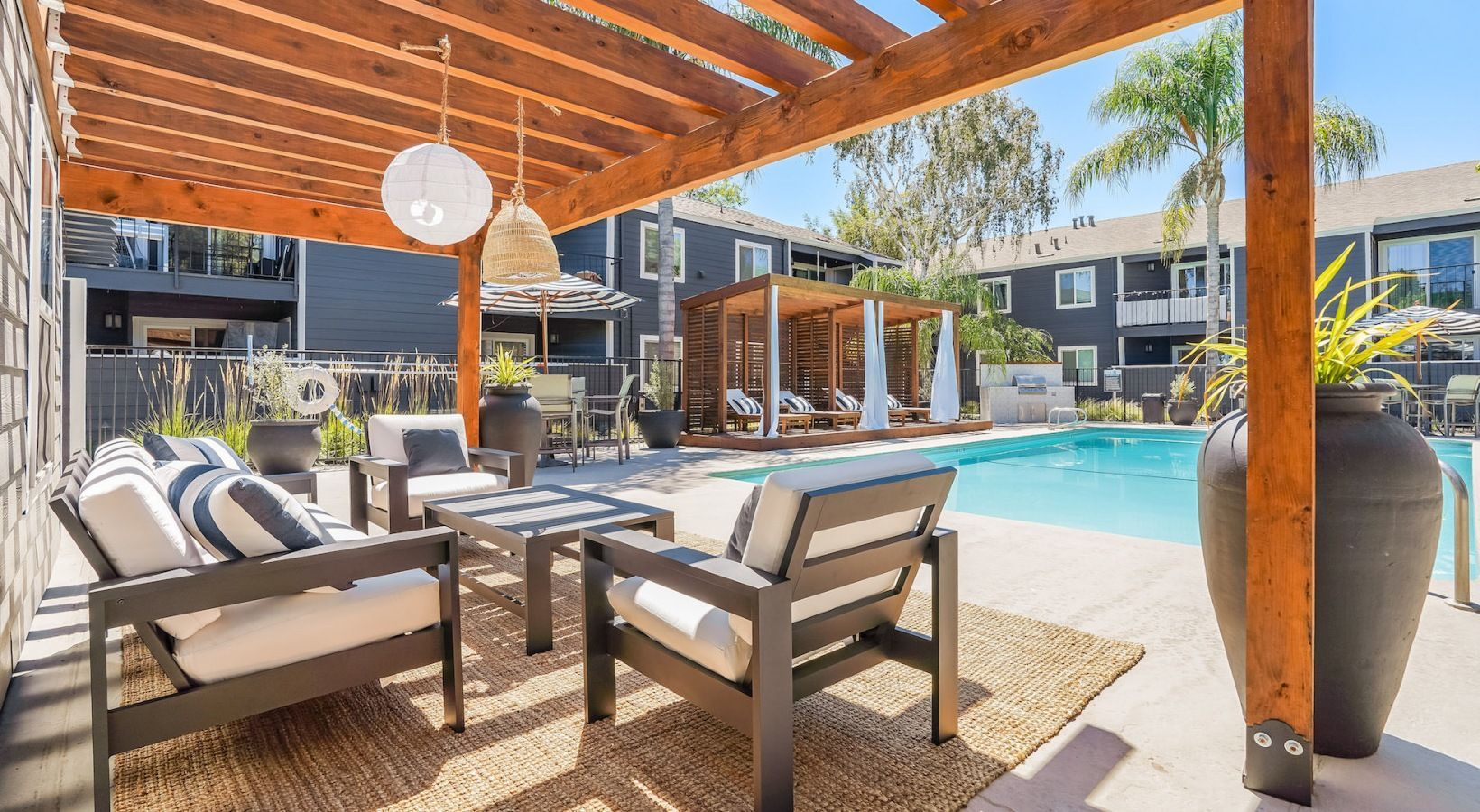 Outdoor pool area with seating under a pergola, near apartments and palm trees.
