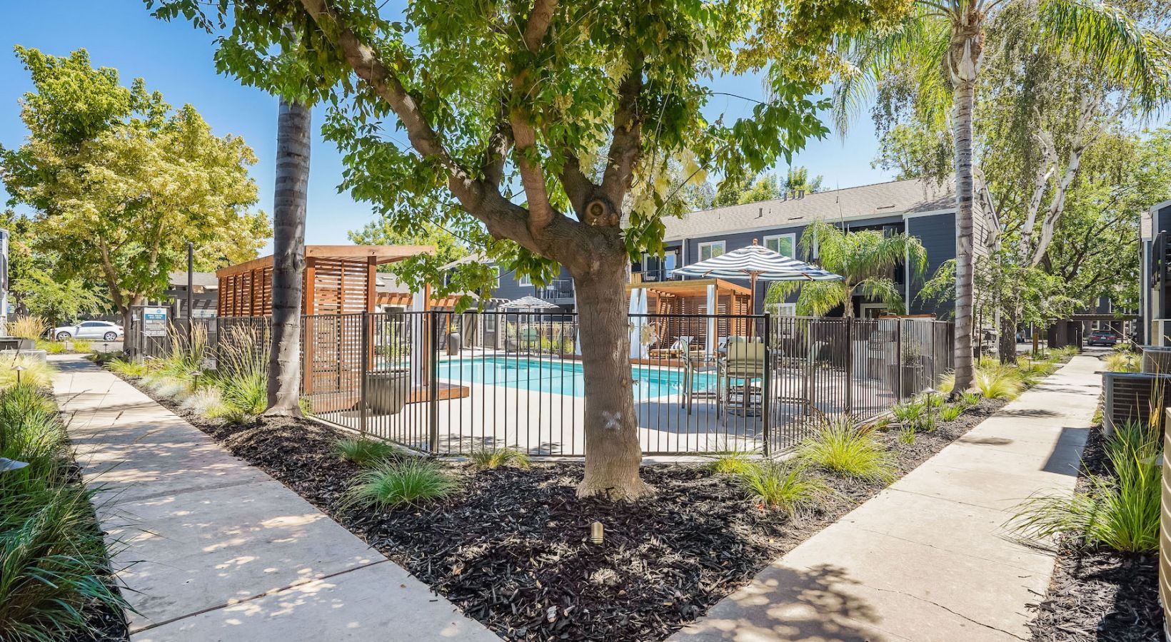 A pool area with fence, trees, and walkway. Sunny day, gray and green tones.