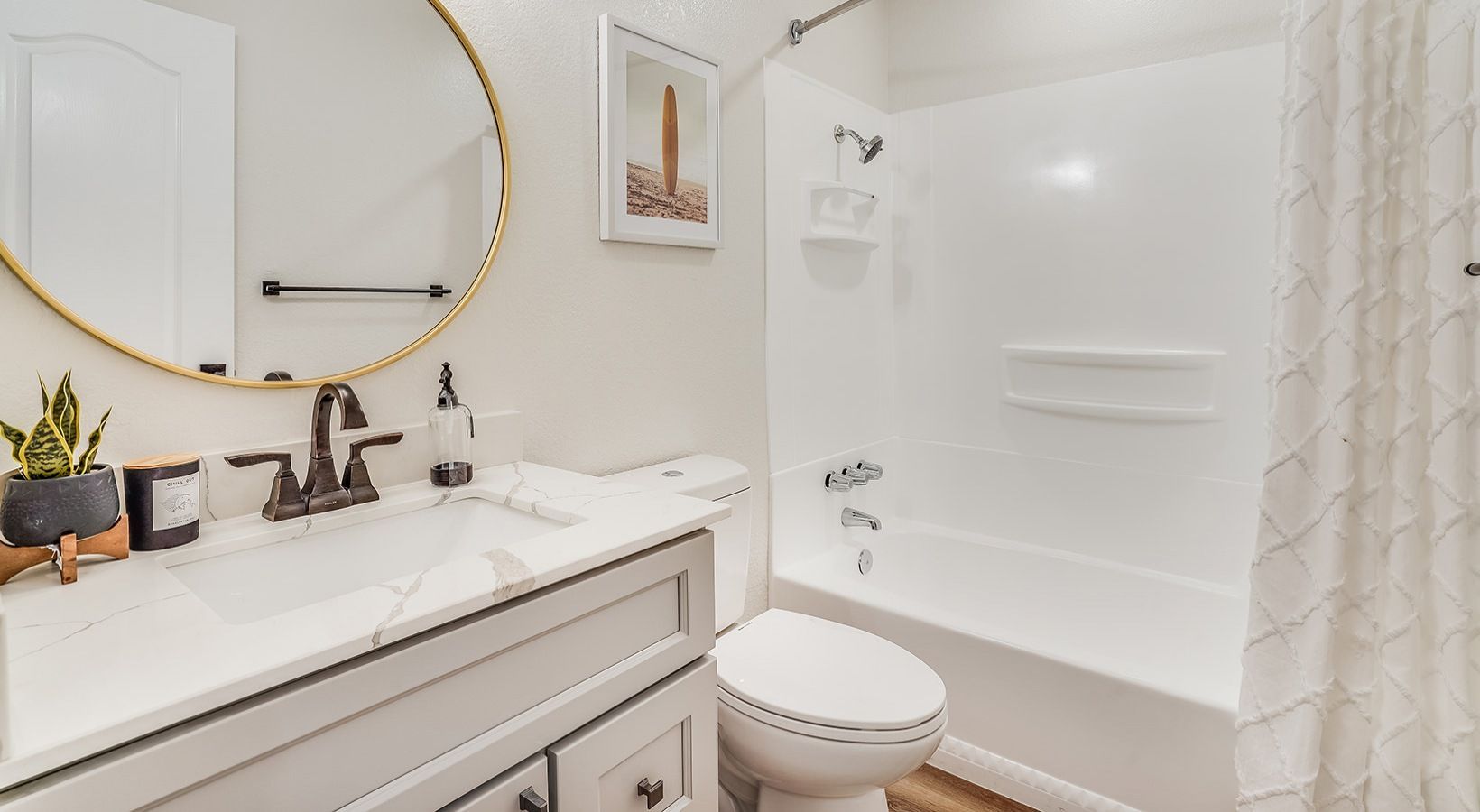 Bathroom with white walls, gray vanity, round gold mirror, and a white bathtub.