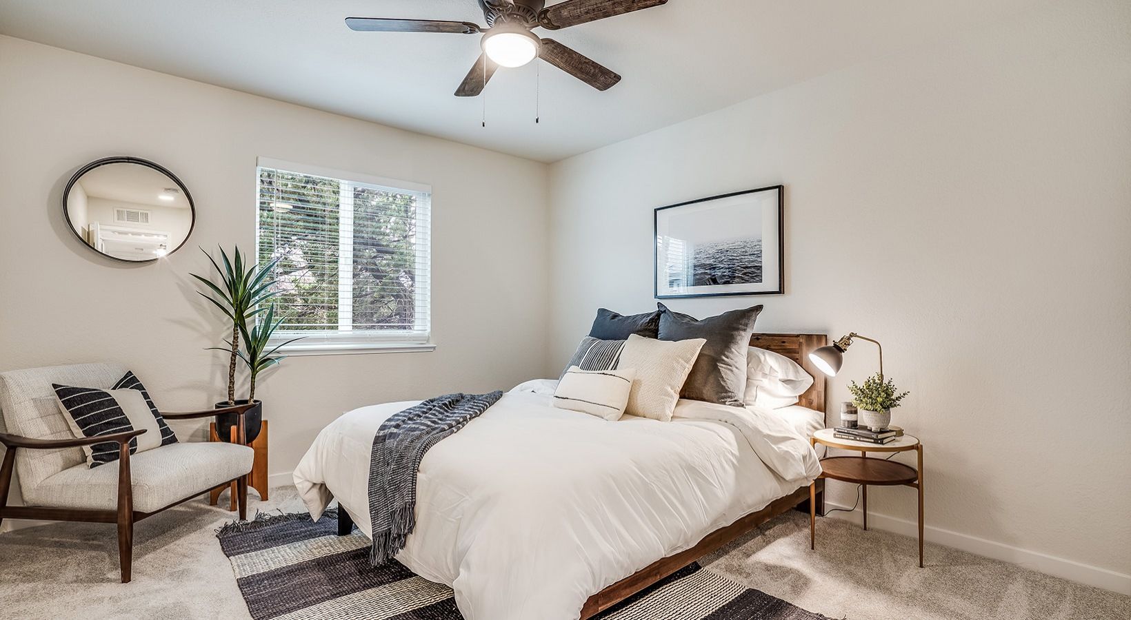 Bedroom with a bed, chair, and artwork; neutral colors, natural light, and a ceiling fan.