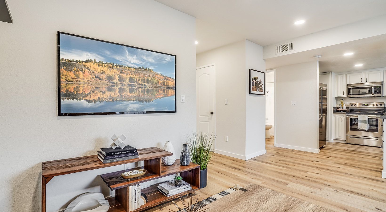 Living room with TV, wooden console, rug, and kitchen visible in the background.
