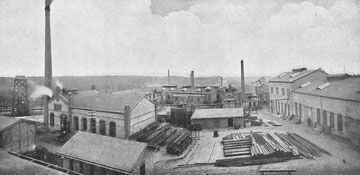 Ein Schwarzweißfoto einer Fabrik, aus deren Schornsteinen Rauch aufsteigt.