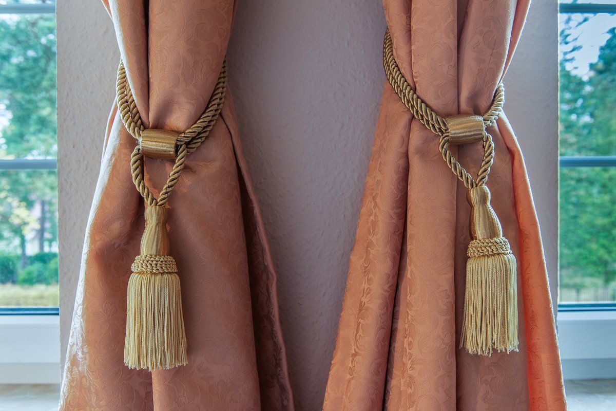 An einem Fenster hängen zwei Vorhänge mit daran befestigten Quasten.