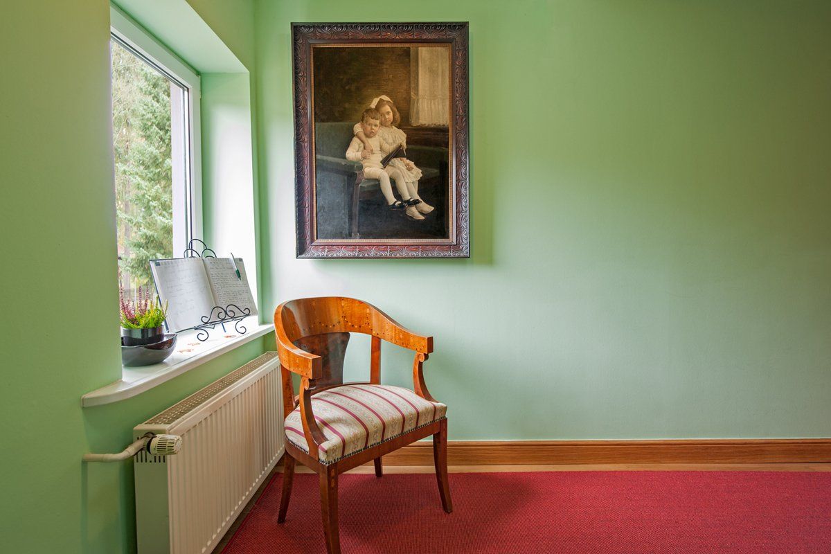 In einem Raum neben einem Fenster steht ein Stuhl, an dessen Wand ein Gemälde hängt.